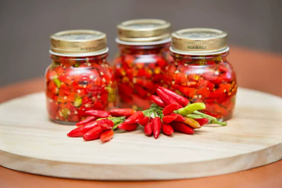 Photo of three jars of canned red Calabrian chili peppers placed on a table. The peppers are from Italy, denoted by the made in Italy label on the lid of the jars. In front of the jars of peppers are a few bunches of Calabrian chili peppers. 