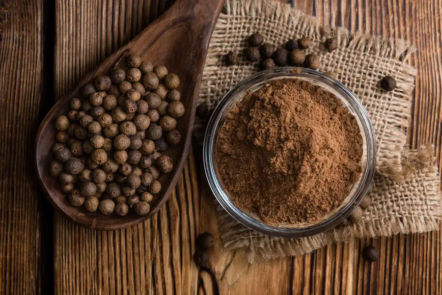 whole Allspice sitting in a large wooden spoon next to ground Allspice in a glass container