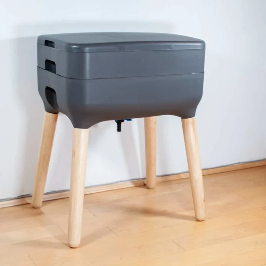Photo of a indoor worm composter standing against white wall. The composter consists of four pine wooden legs with grey curved plastic compartments on top for holding the worms and compost.