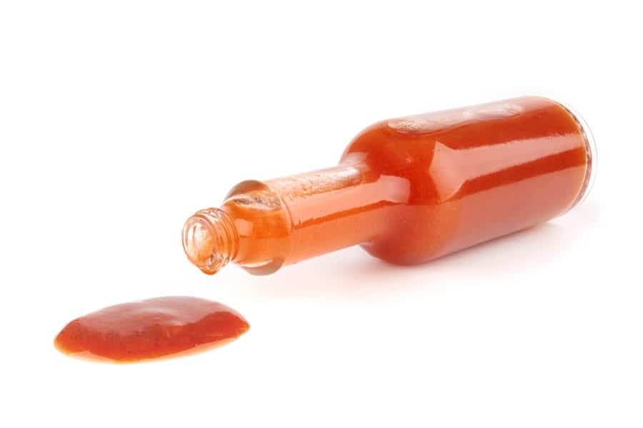 Photo of a tipped over bottle of hot sauce on a white backdrop. The hot sauce has spilt out of the open end of the bottle.