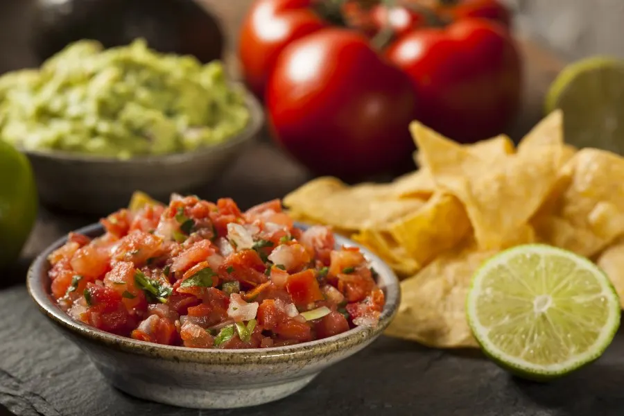Photo of pico de gallo in a medium sized beige ceramic bowl. Behind the pico de gallo is a lime, tortilla chips, guacamole and a batch of tomatoes